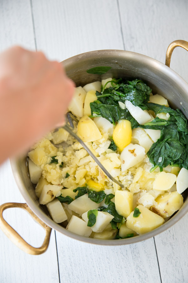 boiled potatoes and spinach in a pot with hand mashing them with potato masher