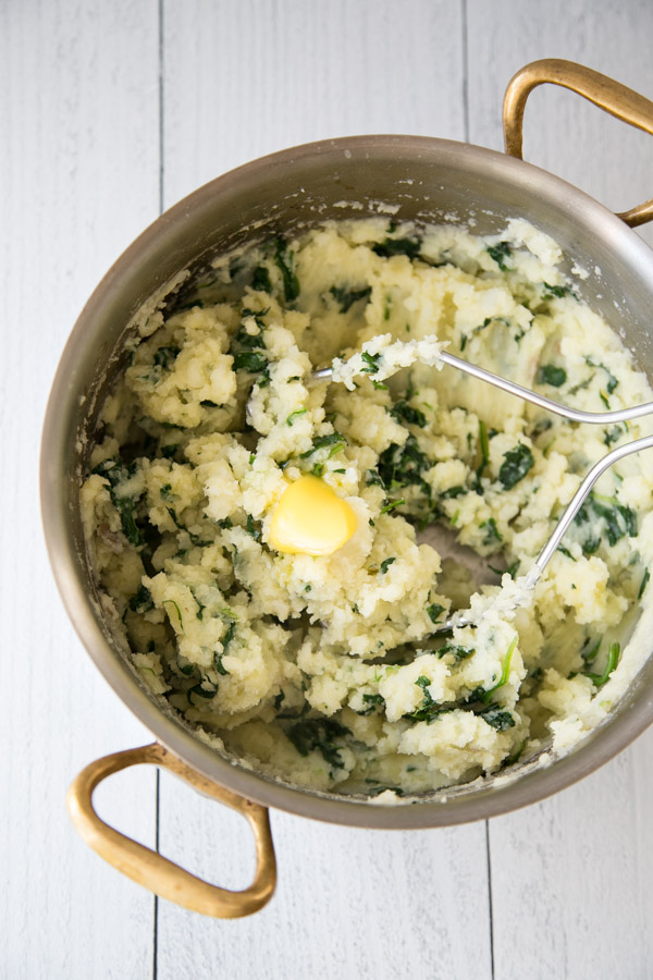 Spinach and potatoes mashed in a saucepan with ghee on top and masher