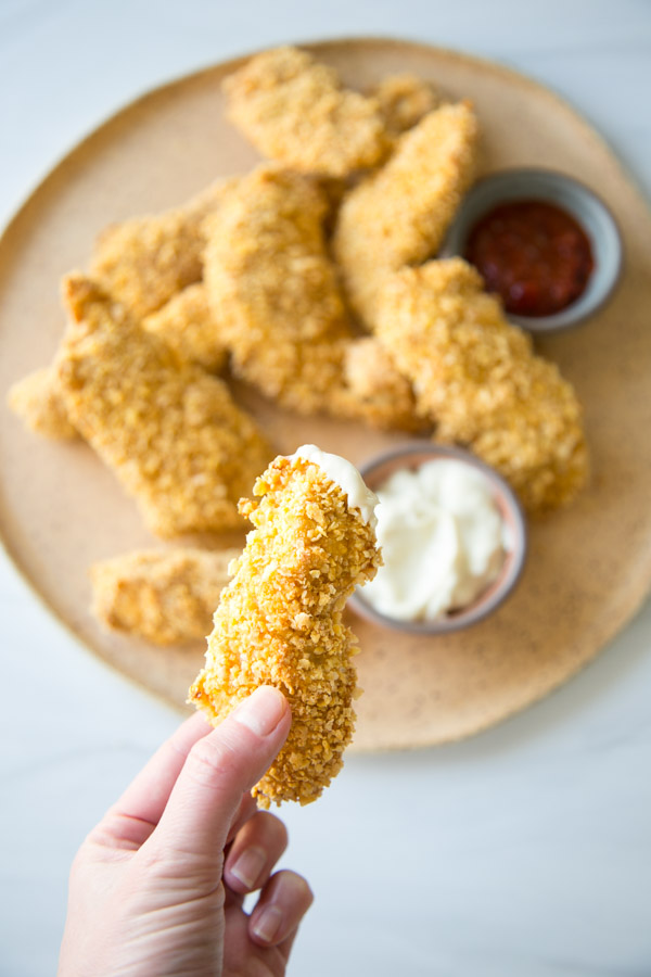 hand holding one crispy baked gluten-free chicken tender over a platter with ketchup and mayo dipping bowls