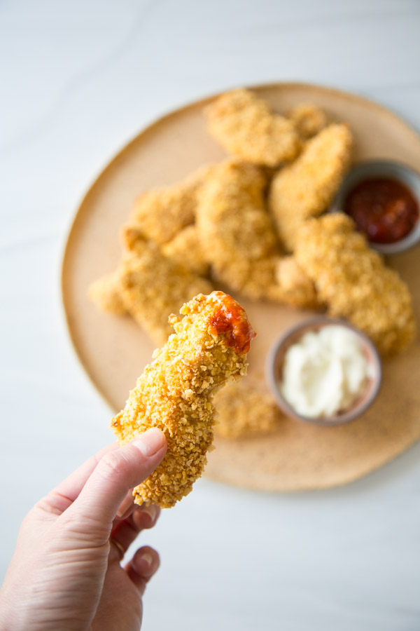 Crispy Baked Gluten-Free Chicken Tenders with Cornflake Crust on a platter. hand holding one chicken strip coated in ketchup