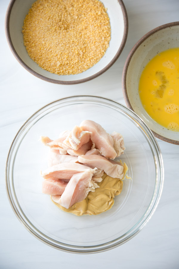 gluten-free cornflake coating in a bowl next to beaten eggs and chicken strips in a third bowl with dijon mustard
