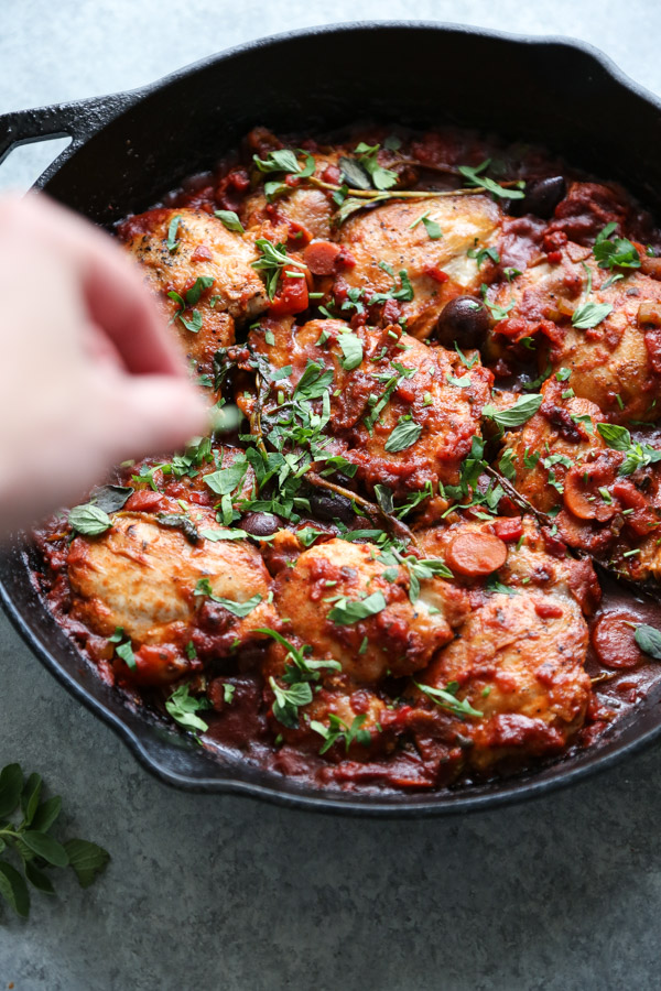 chicken cacciatore in a skillet with oregano and parsley in hand garnishing