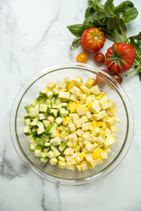 chopped zucchini and summer squash in a mixing bowl next to tomatoes