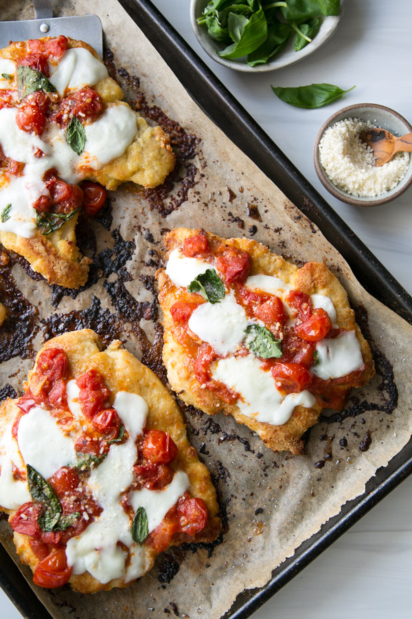 Breaded Crispy Gluten-Free chicken parmesan cutlets topped with fresh tomato sauce and melted mozzarella on a sheet pan with bowl of grated parmesan for garnish
