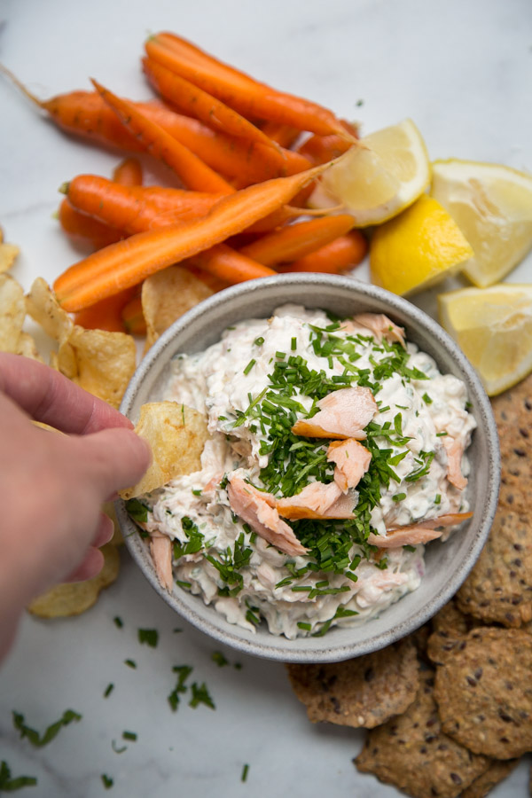 Hand dipping a potato chip into Smoked Salmon Dip in a bowl with chive garnish