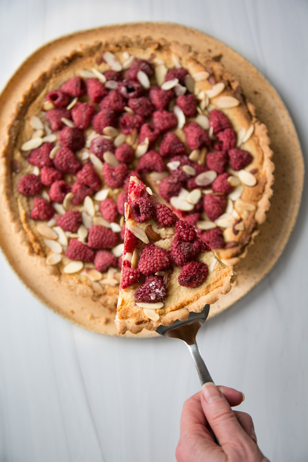 Spatula holding piece of gluten-free raspberry tart on a platter with almonds