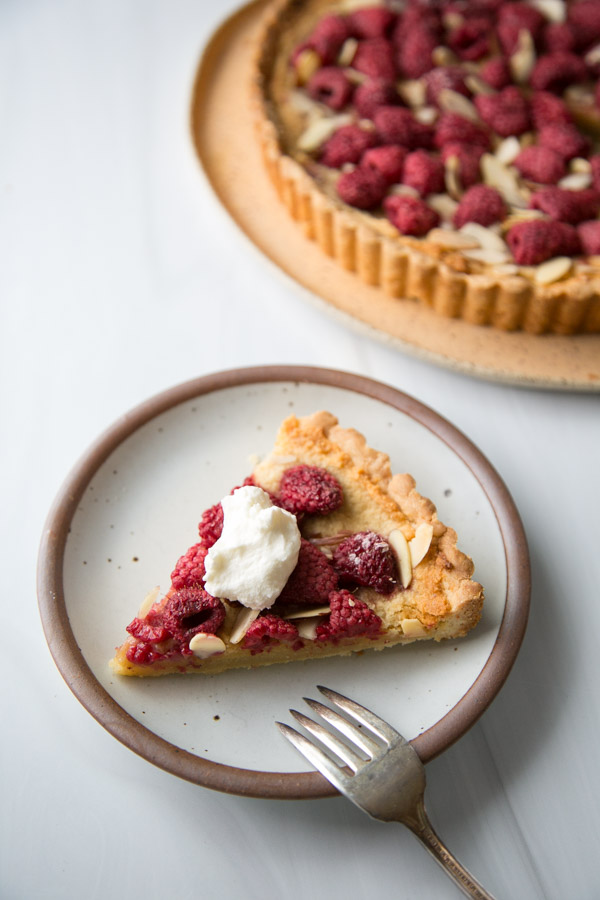 Slice of raspberry frangipan tart on a place with whole almond tart in back
