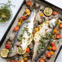 Two whole roasted fish on a baking sheet, garnished with lemon slices, capers, fresh herbs, and surrounded by roasted cherry tomatoes and garlic cloves. A bowl of herb dressing sits nearby.
