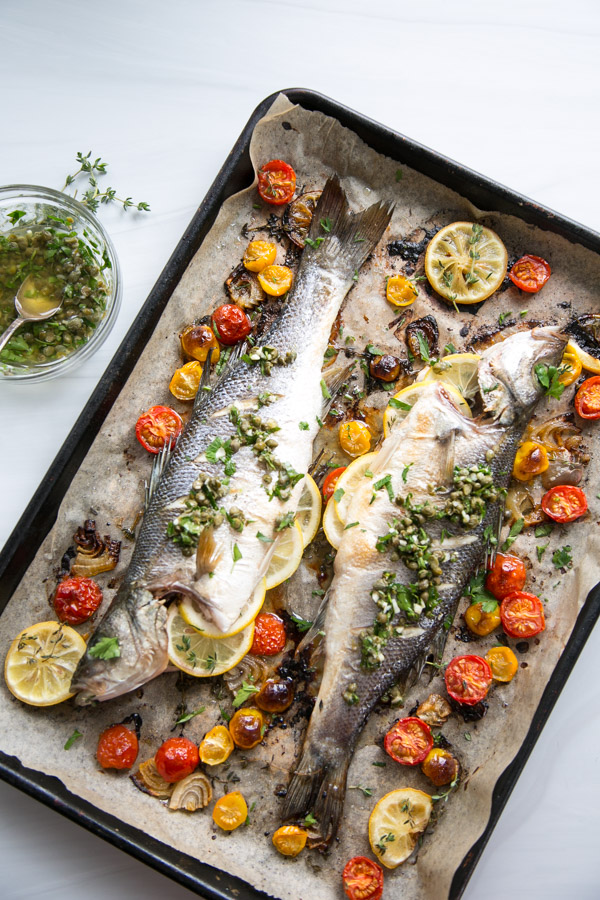 Two whole roasted fish on a baking sheet, garnished with lemon slices, capers, fresh herbs, and surrounded by roasted cherry tomatoes and garlic cloves. A bowl of herb dressing sits nearby.