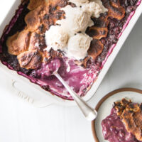 A rectangular baking dish of gluten-free blueberry cobbler with a golden-brown crust, made with almond flour, is topped with vanilla ice cream. A spoon rests inside, and a serving is plated nearby.