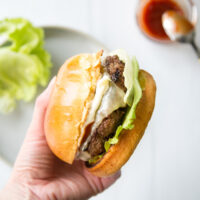 A hand holds up a gluten-free cheeseburger with lettuce and a toasted bun; a plate with lettuce and a jar of ketchup with a spoon are blurred in the background.