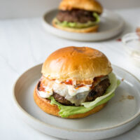 A cheeseburger with lettuce, melted cheese, and sauce sits on a gluten-free brioche bun. Another similar burger is blurred in the background on a separate plate.
