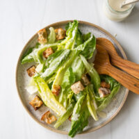 A plate of Caesar salad with romaine lettuce, croutons, and dressing is served with wooden salad tongs. A small glass jar of dressing with a striped straw sits nearby on a white surface.