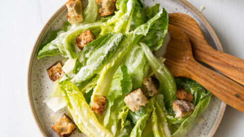 Salade César sans produits laitiers avec vinaigrette maison 20 Un bol de salade César est servi avec de la laitue romaine, des croûtons et de la vinaigrette à l'aide de pinces à salade en bois. Un petit pot en verre contenant de la sauce avec une paille rayée placée près d'une surface blanche.