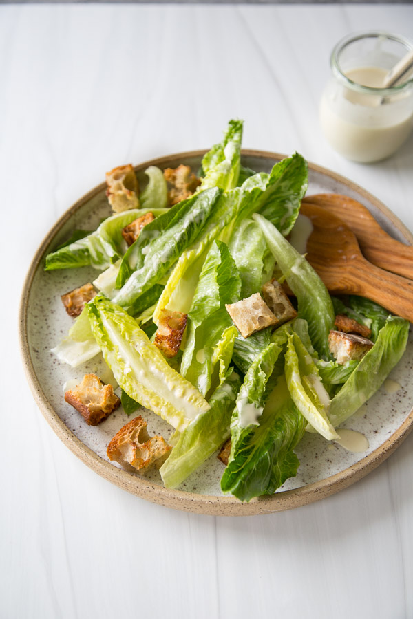 Salade César sans produits laitiers avec vinaigrette maison 14 Une assiette de salade César avec laitue romaine, croûtons et sauce crémeuse, servie avec des ustensiles en bois sur une surface claire. Un petit pot de vêtements apparaît en arrière-plan.