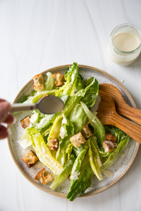 Salade César sans produits laitiers avec vinaigrette maison 12 Un bol de salade César avec de la laitue romaine et des croûtons est arrosé de vinaigrette à la cuillère. Un service à salade en bois et un pot de vinaigrette supplémentaire sont placés près d'un plan de travail blanc.