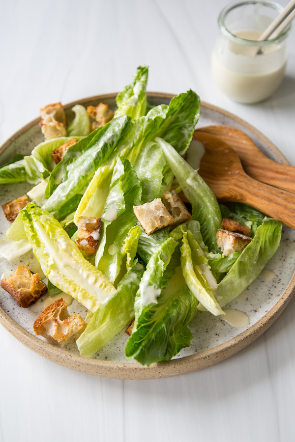 Salade César sans produits laitiers avec vinaigrette maison 18 Assiette de salade César avec laitue romaine, croûtons et vinaigrette crémeuse, servie avec des pinces à salade en bois. Il y a un petit bol de vinaigrettes supplémentaires en arrière-plan sur une surface blanche.