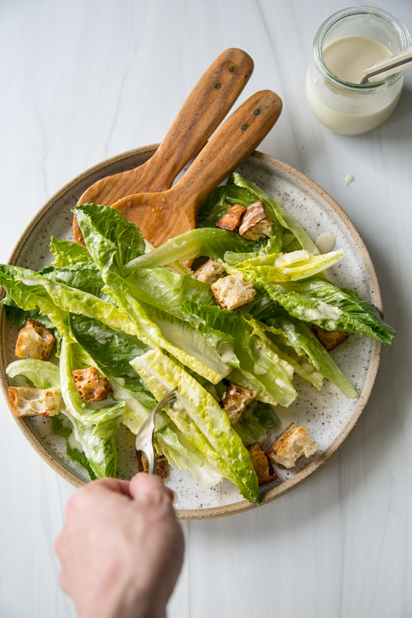 Salade César sans produits laitiers avec vinaigrette maison 16 Fourchette à main, salade César prête à manger avec laitue romaine, croûtons et vinaigrette sur une assiette avec une salade en bois ; Un bol de vinaigrettes supplémentaires se trouve près d’une surface blanche.