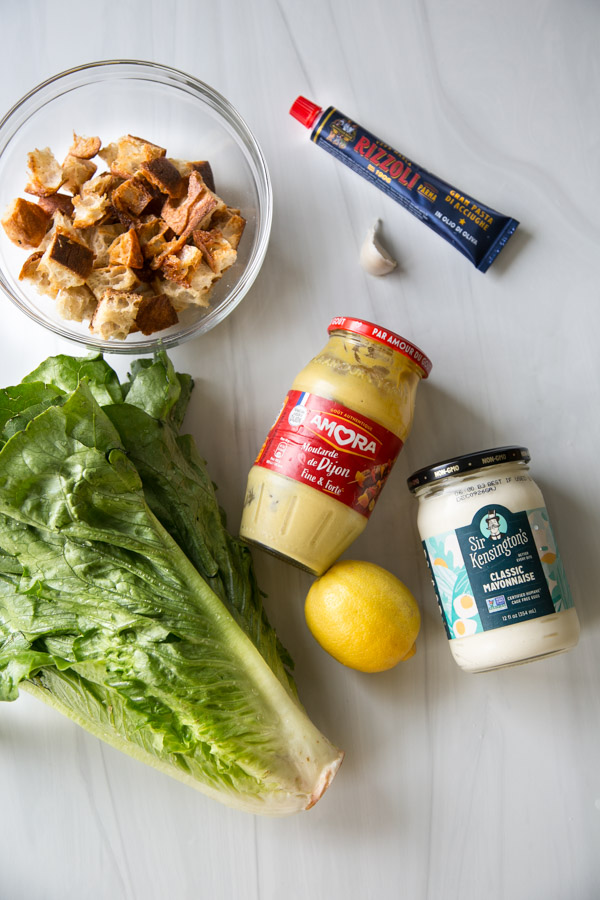 Salade César sans produits laitiers avec vinaigrette maison 2 Un bol de cubes de pain, un tube de pâte d'anchois, une gousse d'ail, de la moutarde de Dijon, un pot de mayonnaise, un citron et une tête de laitue romaine sont disposés sur une surface blanche.