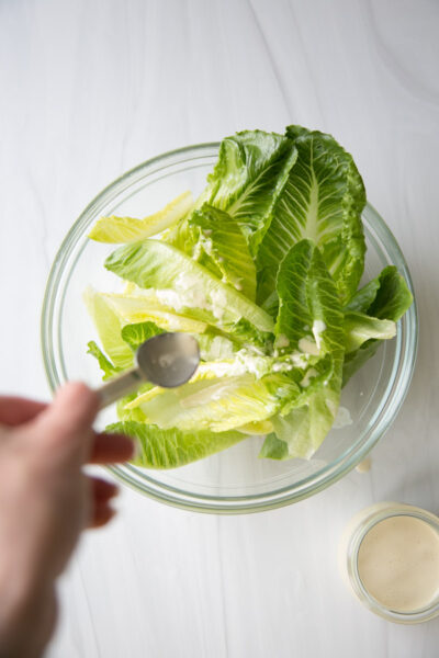Salade César sans produits laitiers avec vinaigrette maison 8 Une main tenant une cuillère verse une sauce crémeuse sur des feuilles de laitue romaine fraîches dans un bol en verre transparent, avec un petit pot de sauce près de la surface blanche.