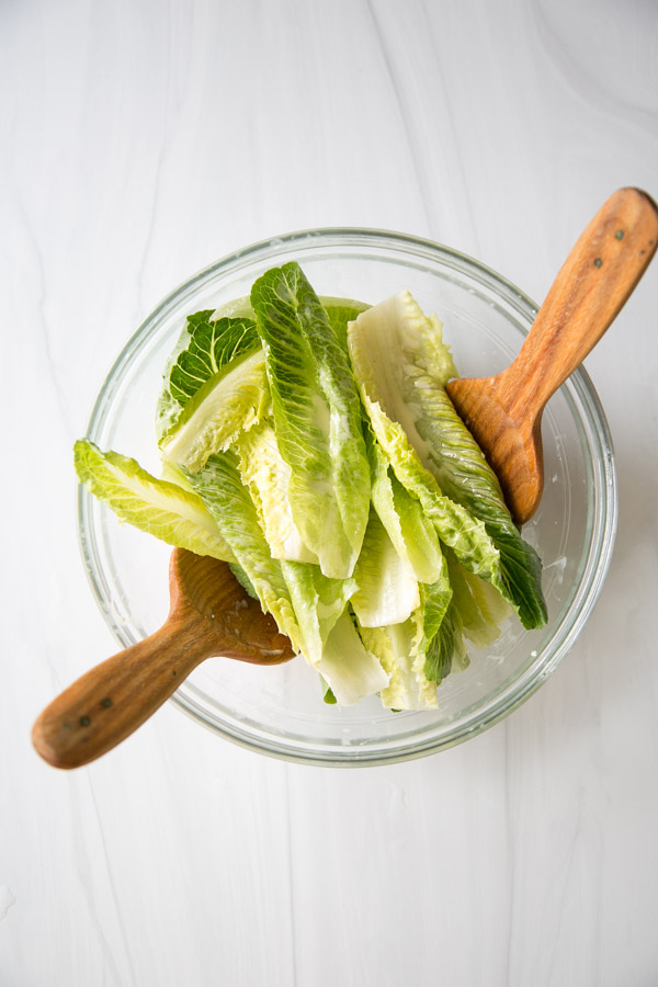 Salade César sans produits laitiers avec vinaigrette maison 10 Un bol en verre rempli de feuilles de laitue romaine fraîche, avec deux assiettes à salade en bois sur les côtés, vues de dessus sur une surface blanche.