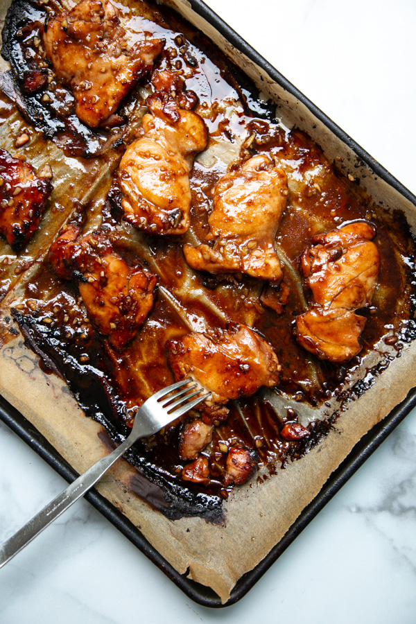 Baked, glazed chicken thighs on a parchment-lined baking tray, with a fork resting on the tray. The chicken appears caramelized and saucy, against a white marble background.