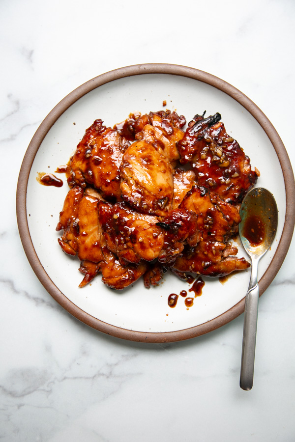 A plate of glazed, saucy chicken pieces garnished with herbs, with a spoon resting on the rim, on a white marble surface.