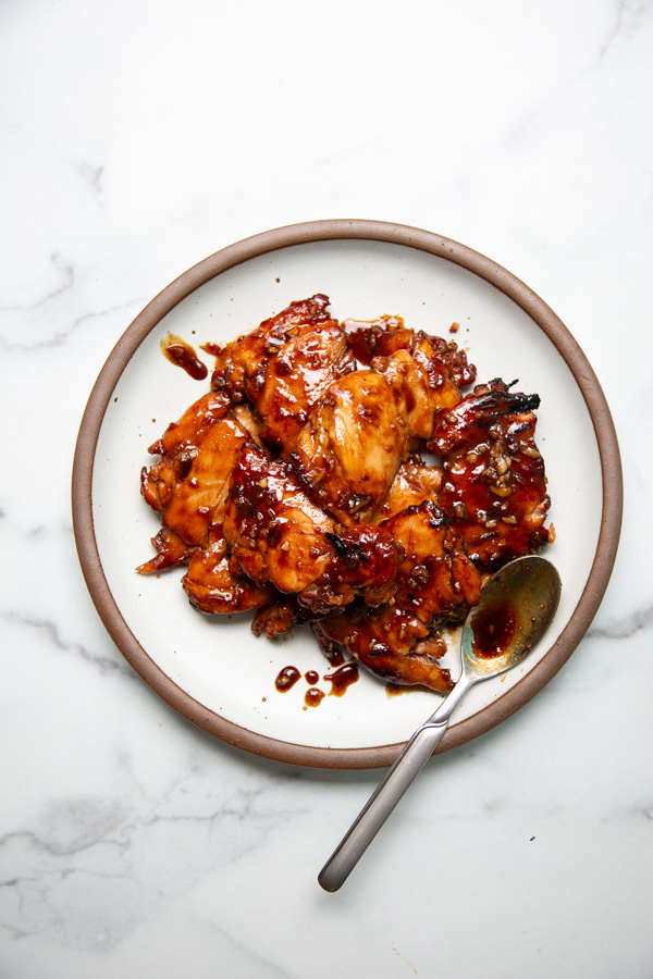 A plate of glazed chicken wings coated in a dark, sticky sauce with a spoon resting on the side, set on a white marble surface.