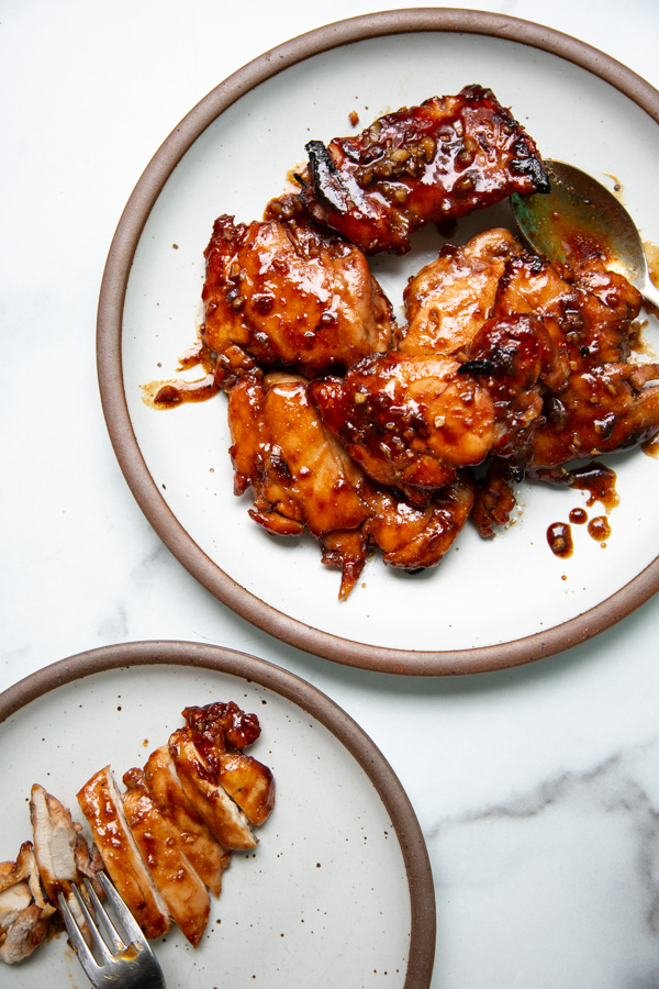 Two plates with glazed, cooked chicken; the larger plate holds several pieces coated in a dark sauce, while the smaller plate has sliced chicken with sauce and a fork on the side. Both plates have brown rims and rest on a white surface.