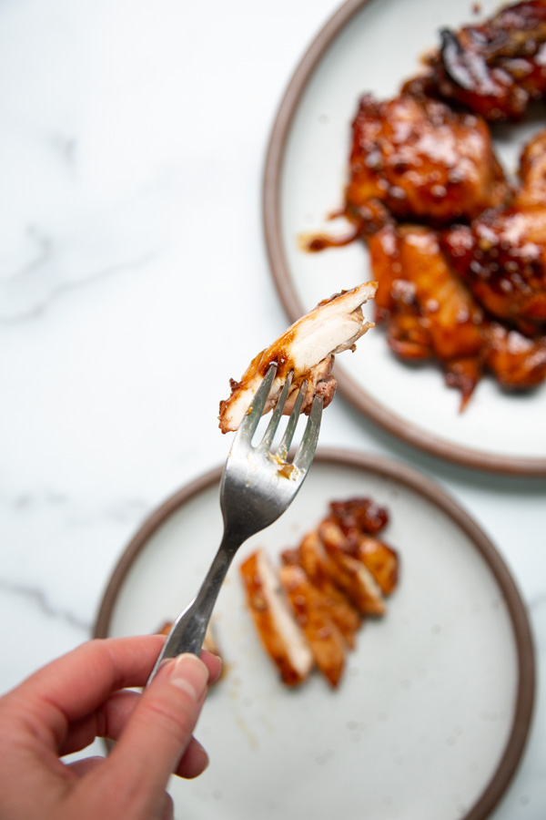 A hand holds a fork with a piece of glazed cooked chicken, with plated pieces of chicken on two blurred plates in the background.