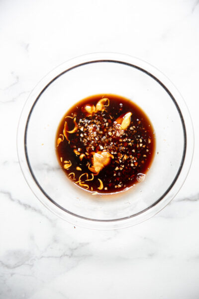 A glass bowl on a white marble surface filled with a dark liquid marinade containing chopped garlic, onion, and orange peel.