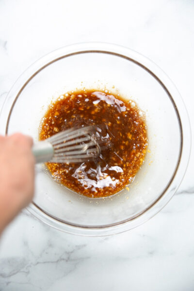 A hand uses a metal whisk to mix a brown sauce with visible bits in a clear glass bowl on a white marble surface.