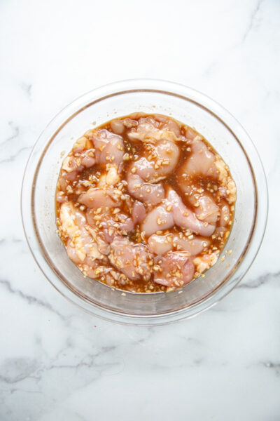 Raw chicken pieces marinating in a glass bowl with minced garlic and a brown sauce, placed on a white marble surface.