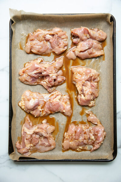Raw marinated chicken thighs spread out on a parchment-lined baking sheet, ready to be cooked. The baking sheet is placed on a white marble surface.