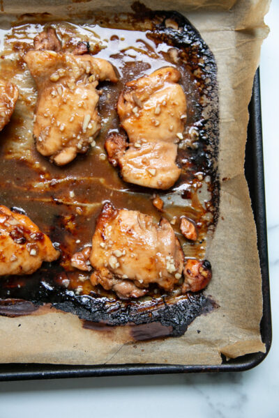 Baked chicken thighs coated in a glossy garlic marinade sit on parchment paper atop a baking tray; some sauce has caramelized around the edges.