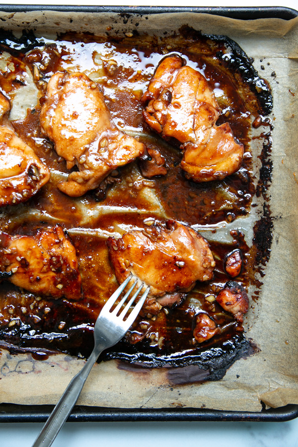 Baked chicken thighs coated in a glossy brown sauce on a parchment-lined baking sheet, with a fork resting near the chicken.