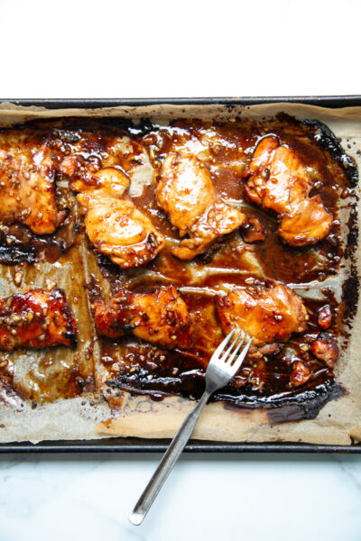 Baked chicken thighs glazed with a dark, sticky sauce on a parchment-lined baking sheet, with a silver fork resting on the tray.