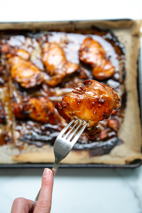 A hand holds a fork with a glazed piece of baked chicken above a tray lined with parchment paper, featuring more glazed chicken pieces in the background.