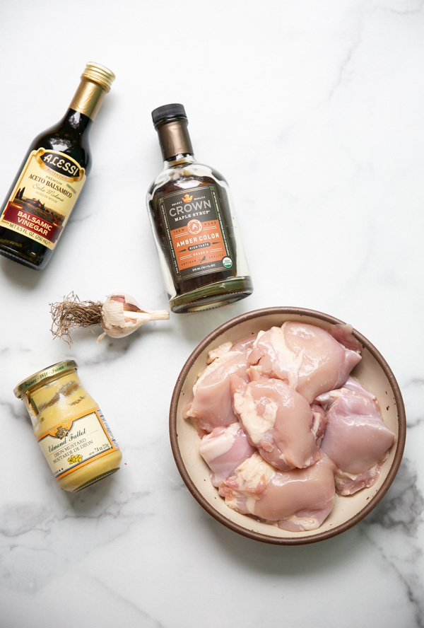 A bowl of raw chicken thighs, a bottle of balsamic vinegar, a bottle of maple syrup, a jar of Dijon mustard, and a garlic bulb on a white surface.