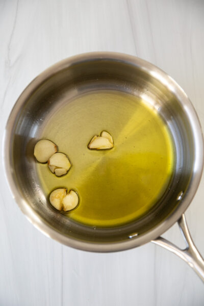 A stainless steel saucepan with olive oil and a few slices of garlic on the bottom, sitting on a white marble surface.