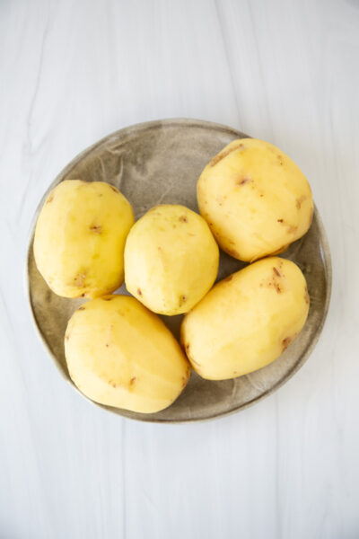 Five peeled potatoes sit on a round beige plate, placed on a white marble surface.