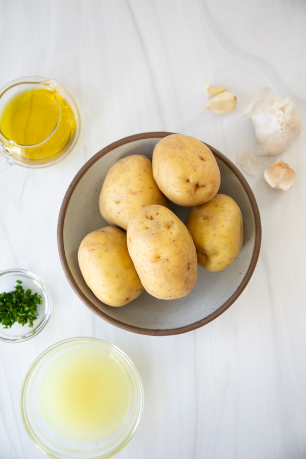 A bowl of whole potatoes surrounded by a small jar of olive oil, garlic cloves, chopped herbs, and a bowl of broth on a white surface.