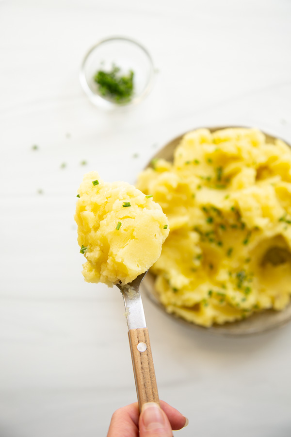 A hand holds a spoonful of mashed potatoes topped with chopped chives, with a plate of mashed potatoes and a small bowl of chives in the background.