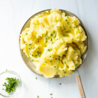 A plate of mashed potatoes topped with melted butter and chopped chives, with a small bowl of chives on the side. A wooden-handled spoon rests on the plate.