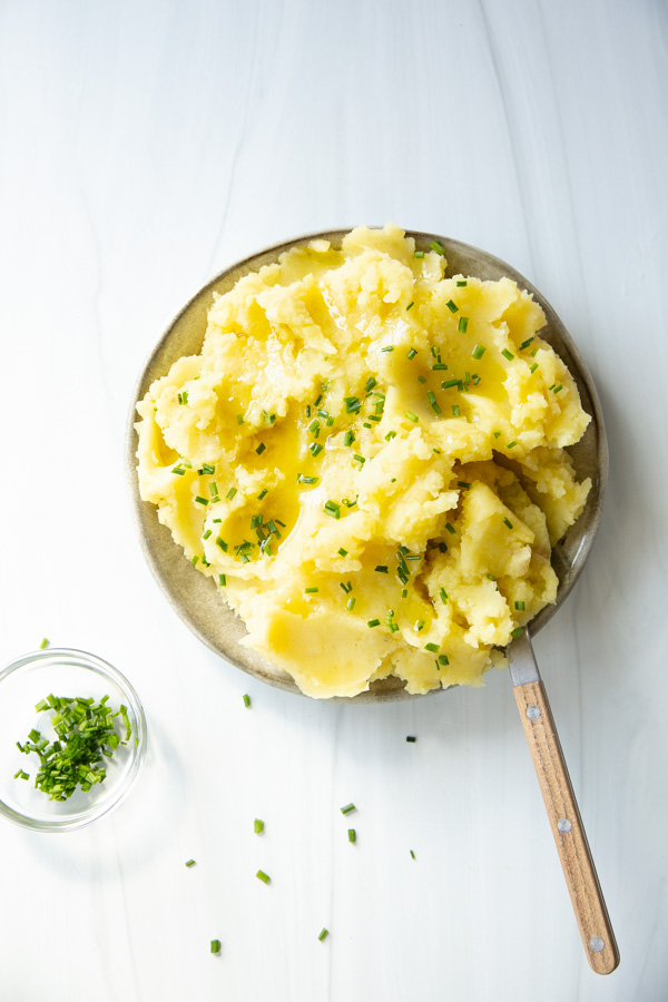 A plate of mashed potatoes topped with melted butter and chopped chives, with a small bowl of chives on the side. A wooden-handled spoon rests on the plate.