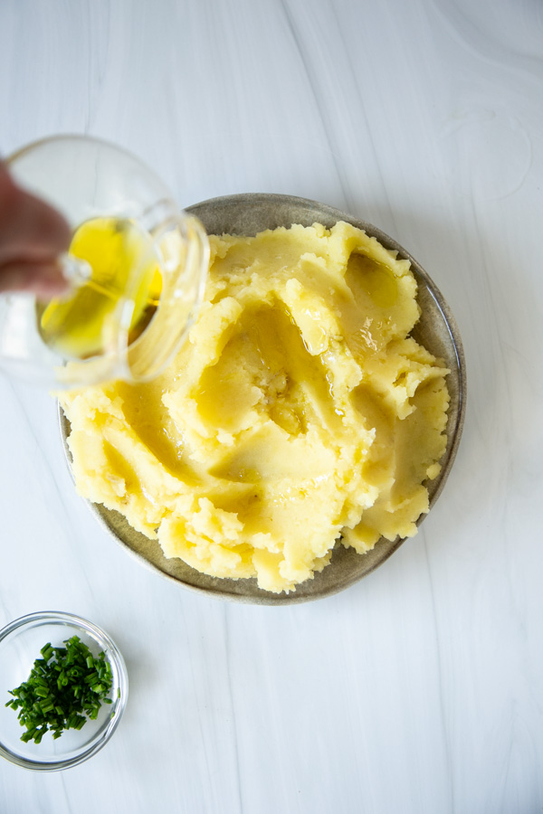 A hand pours olive oil onto a plate of mashed potatoes. A small bowl of chopped green herbs sits to the side on a white surface.