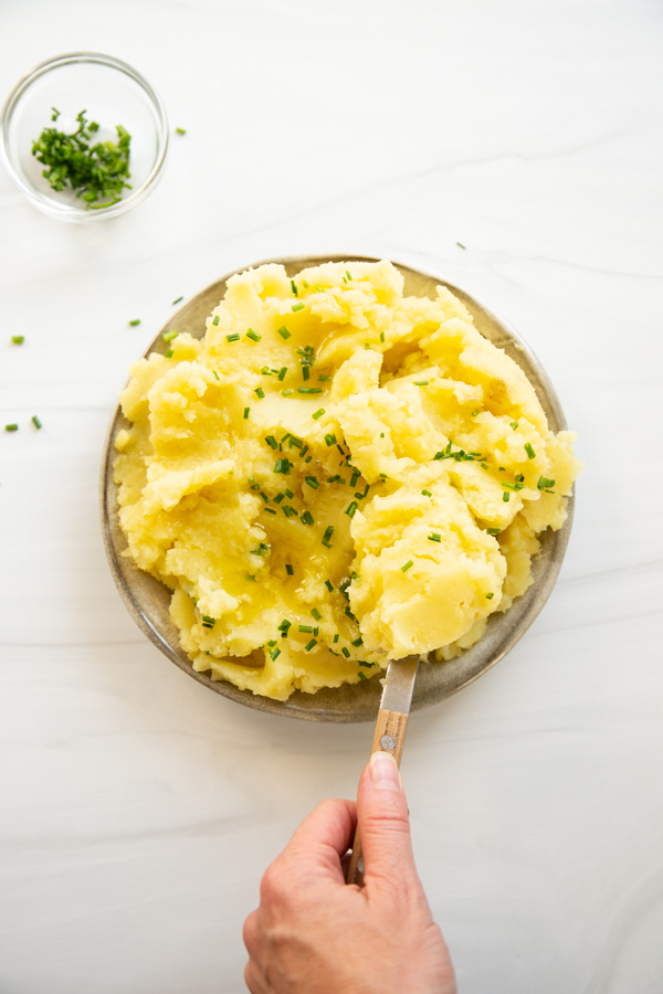 A hand holds a fork, scooping creamy mashed potatoes garnished with chopped chives on a gray plate. A small bowl of extra chives sits nearby on a white surface.