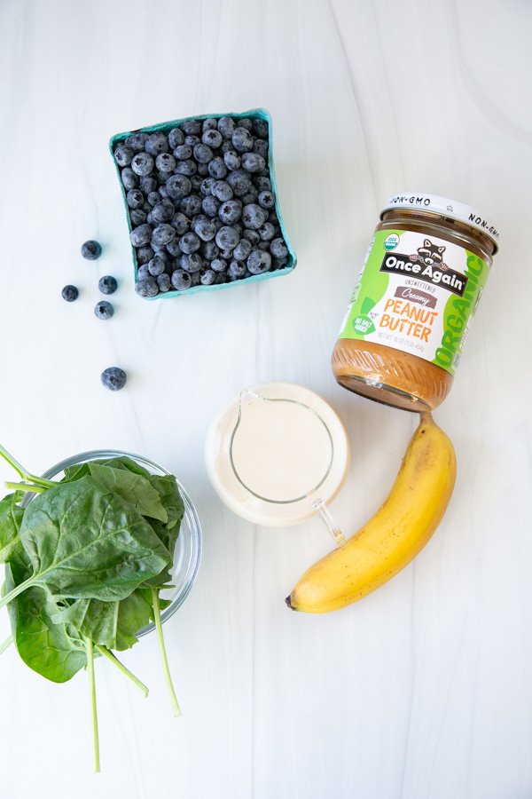 A glass of spinach, a carton of blueberries, a banana, a jar of peanut butter, and a small glass pitcher of milk are arranged on a white surface.