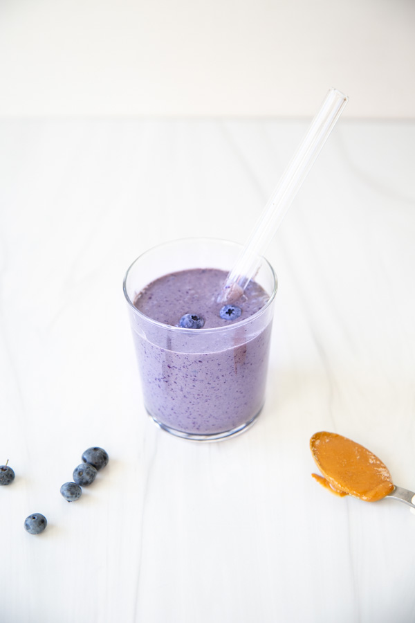 A glass of blueberry smoothie with two blueberries on top and a clear straw, next to scattered blueberries and a spoonful of peanut butter on a white surface.