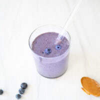A glass of purple blueberry smoothie with a straw, topped with blueberries. Nearby, there are scattered blueberries and a spoon with peanut butter on a white surface.